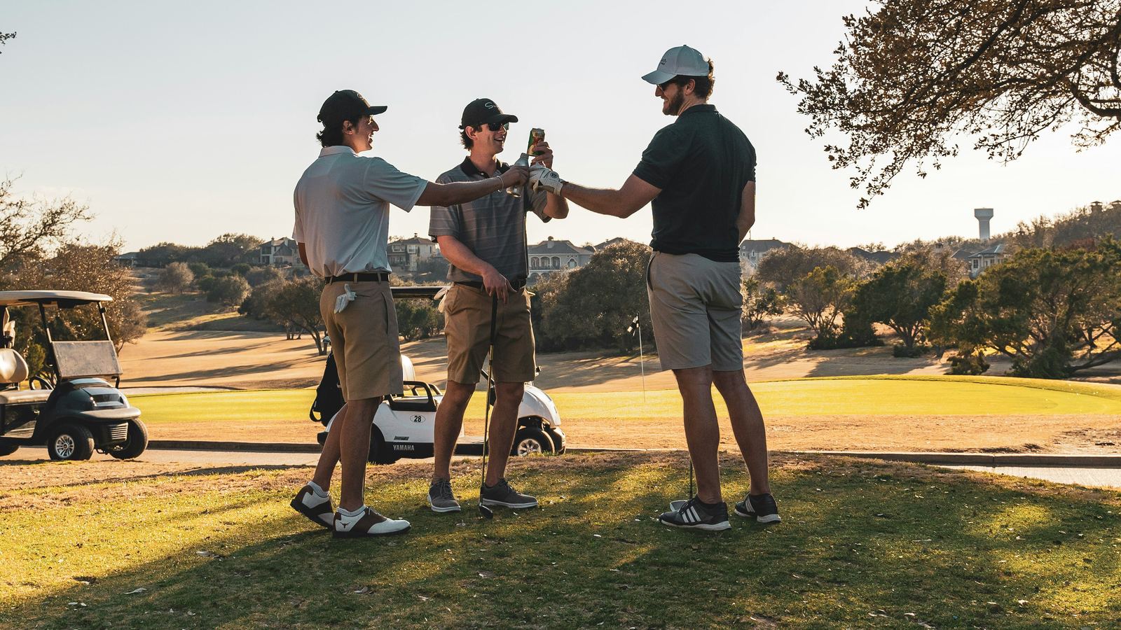 Three golfers on the course at dusk, raising drinks together after a round.