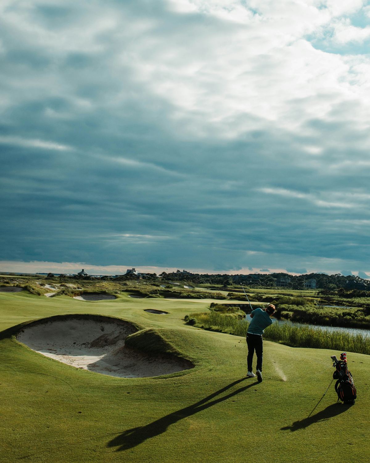 A single player at the top of a swing on a links course, heavy cloud above and a bag standing behind them.