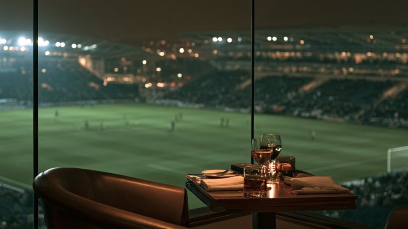 A private box at a stadium, a table set by the glass, the pitch lit up beyond it.